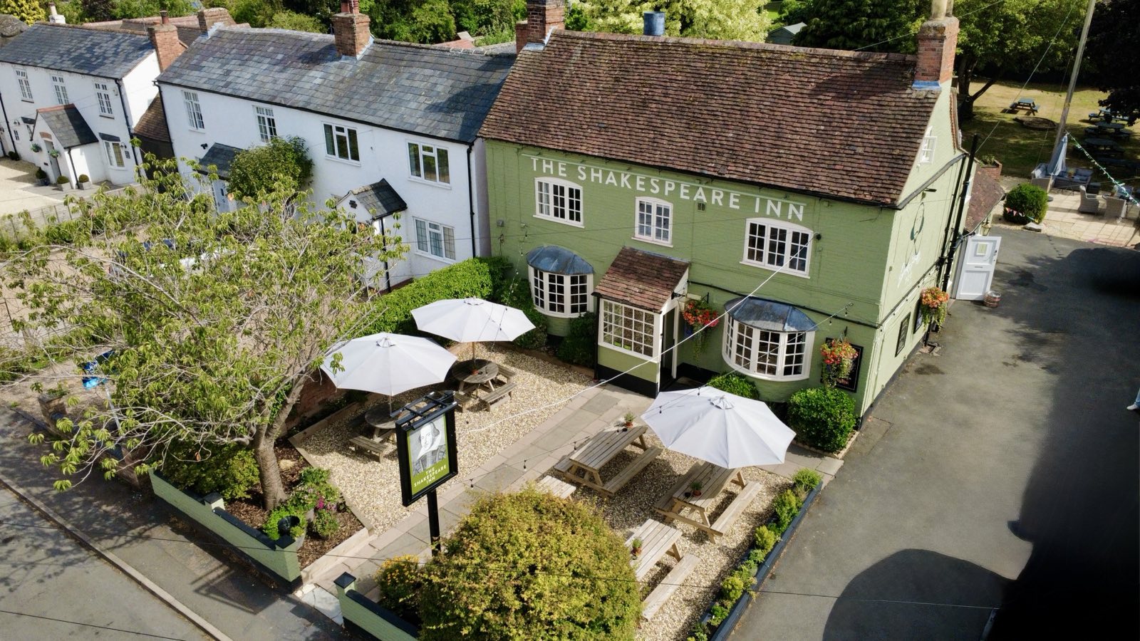 Aerial view of The Shakespeare Inn's front garden - gravel, picnic benches and white parasols in front of the sage-green pub.