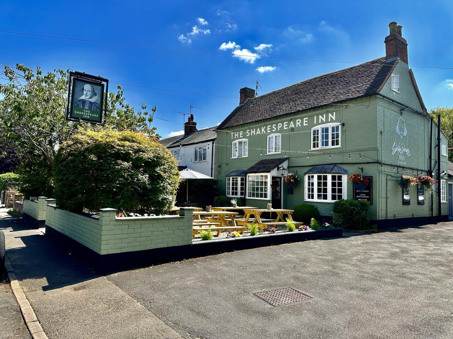 The Shakespeare Inn's sage-green frontage under a blue sky, with the hanging pub sign and picnic benches out front.